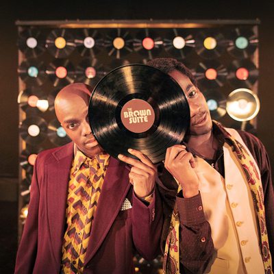 Two Black men in colorful retro-inspired outfits pose in front of a wall of illuminated circular lights. The man in front holds a large vinyl record labeled “The Brown Suite” over his face, while the other man stands behind him, smiling toward the camera.