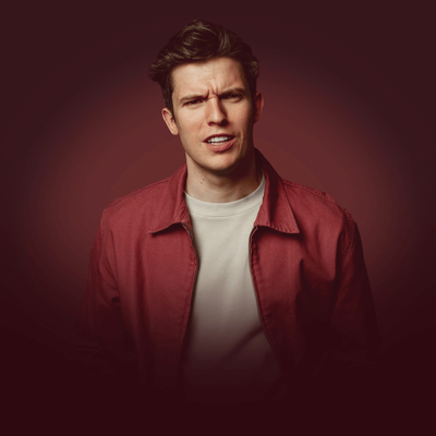 Portrait of a Patrick Spicer with dark, swept-back hair wearing a red jacket over a light-colored shirt, looking toward the camera with a serious expression against a dark burgundy background.