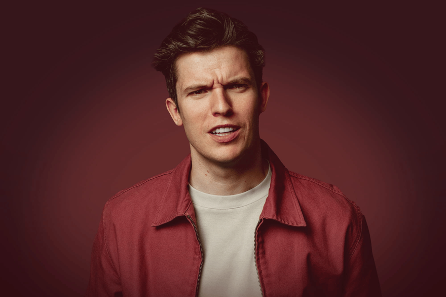 Portrait of a Patrick Spicer with dark, swept-back hair wearing a red jacket over a light-colored shirt, looking toward the camera with a serious expression against a dark burgundy background.
