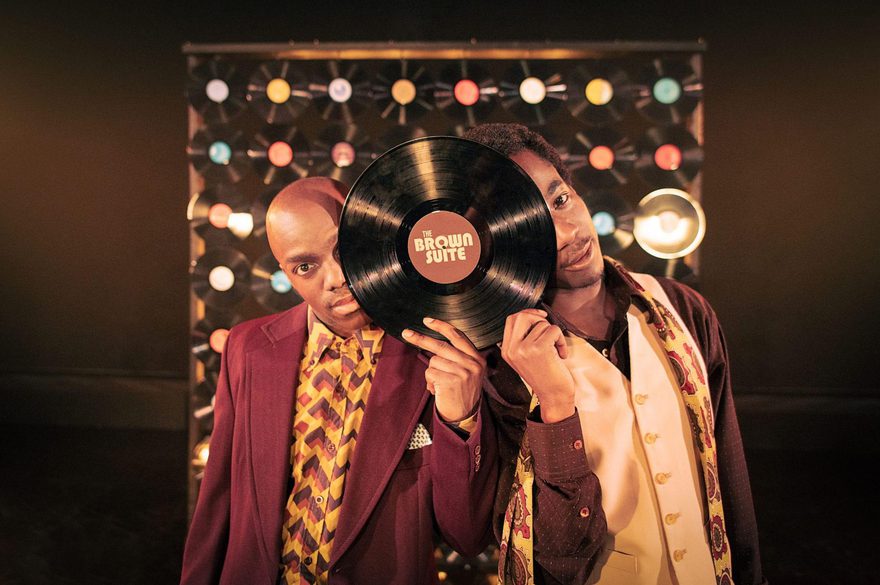 Two Black men in colorful retro-inspired outfits pose in front of a wall of illuminated circular lights. The man in front holds a large vinyl record labeled “The Brown Suite” over his face, while the other man stands behind him, smiling toward the camera.