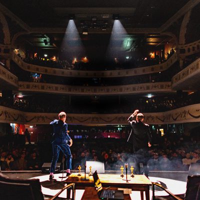 Two performers stand onstage facing a large audience inside an ornate, multi-level theater. One performer raises a hand while the other stands nearby, with stage lights and haze creating a dramatic atmosphere. Ornamental balconies filled with spectators surround the venue.