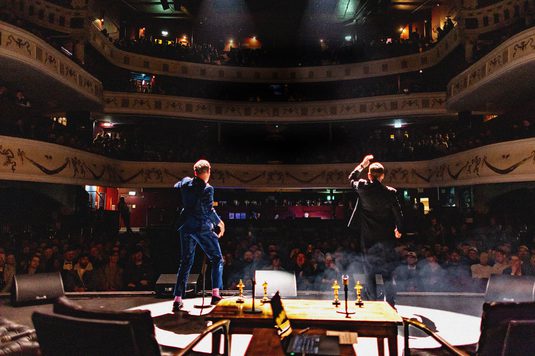 Two performers stand onstage facing a large audience inside an ornate, multi-level theater. One performer raises a hand while the other stands nearby, with stage lights and haze creating a dramatic atmosphere. Ornamental balconies filled with spectators surround the venue.