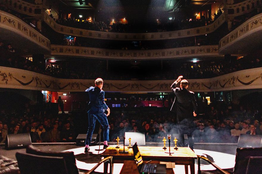 Two performers stand onstage facing a large audience inside an ornate, multi-level theater. One performer raises a hand while the other stands nearby, with stage lights and haze creating a dramatic atmosphere. Ornamental balconies filled with spectators surround the venue.