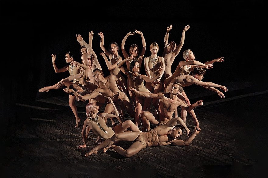 A large group of dancers pose in a dramatic, tightly arranged formation on a dark stage, with several dancers extending their arms and legs in different directions and one dancer reclining across the foreground.