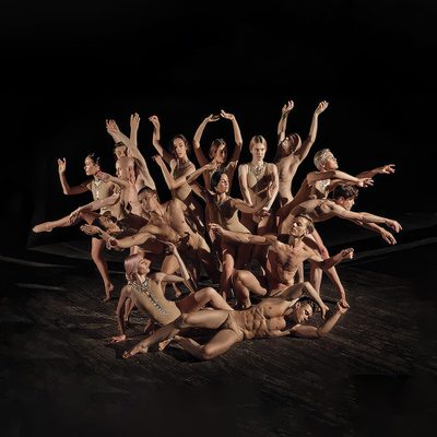 A large group of dancers pose in a dramatic, tightly arranged formation on a dark stage, with several dancers extending their arms and legs in different directions and one dancer reclining across the foreground.