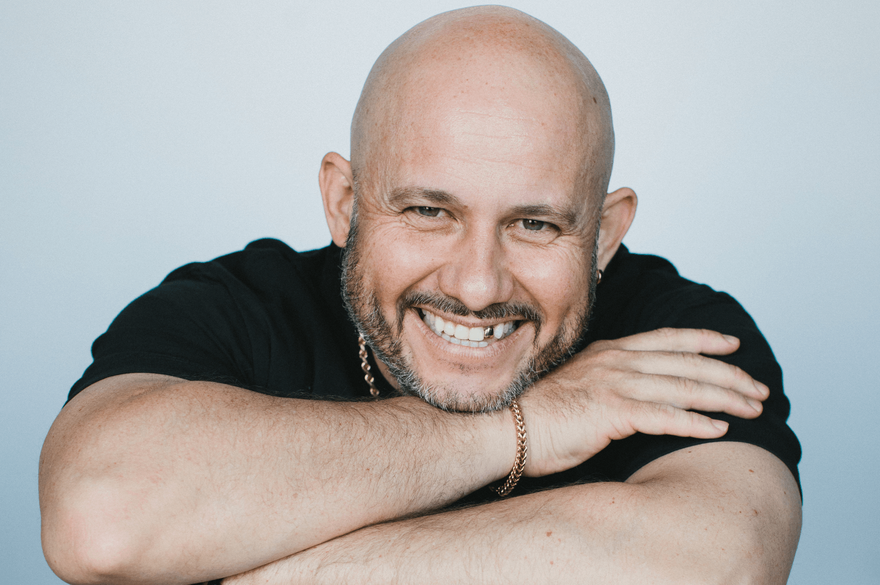 White Yardie Smiling bald man with is a bold white man with a beard, wearing a black shirt, leaning forward with his arms crossed over the back of a black chair.