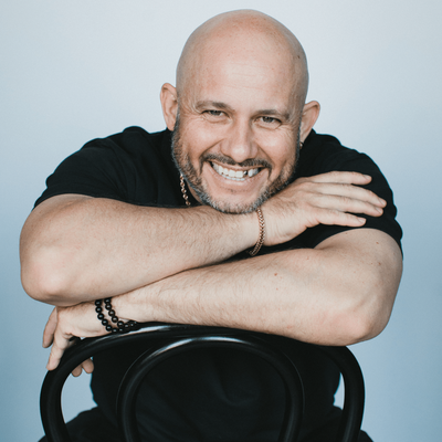 White Yardie Smiling bald man with is a bold white man with a beard, wearing a black shirt, leaning forward with his arms crossed over the back of a black chair.