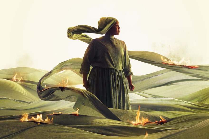 A woman in a green dress stands gracefully in a vibrant field, surrounded by tall grass and wildflowers and little fires.