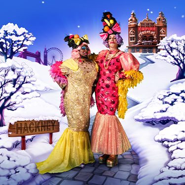 Kat B and George Heyworth as the Ugly Sisters wearing yellow and pink dresses with fruit headdresses. They pose against a snowy scene with Hackney Empire in the background.