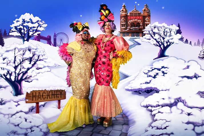 Kat B and George Heyworth as the Ugly Sisters wearing yellow and pink dresses with fruit headdresses. They pose against a snowy scene with Hackney Empire in the background.