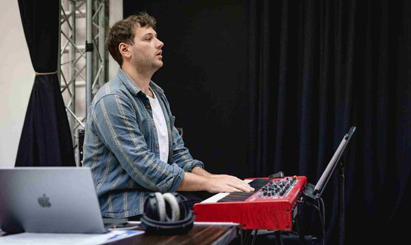 Alex Maynard is playing on the keyboard and looking straight out at the performers.