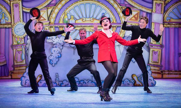 Alexandra Waite Roberts as Oblivia is wearing a red blazer jacket singing with arms outstretched and the Ensemble pose around her with shocked expressions and their hats off.