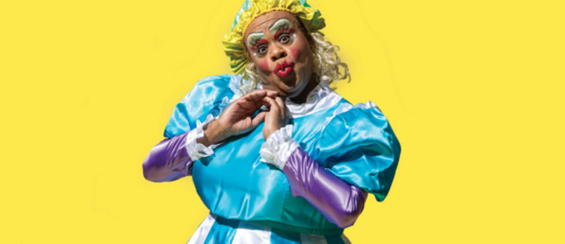 A photo of Clive Rowe dressed in a blue and white dress with a yellow wig and their hands held up to their face.