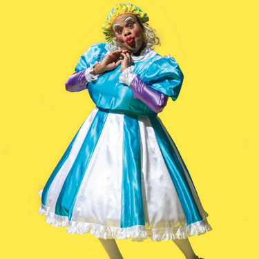 A photo of Clive Rowe dressed in a blue and white dress with a yellow wig and their hands held up to their face.