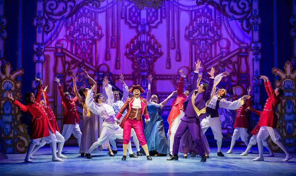 Chrissie Bhima as Dandini is singing whilst Luke Latchman as Prince Charming and the Ensemble dancing with arms up in the air.