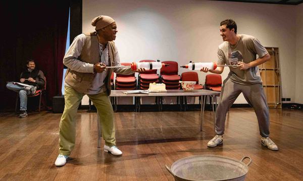 Clive Rowe as Sarah the Cook and Max Mirza as Idle Jack are facing each other holding devices that look like water pistols with gleeful expressions on their faces