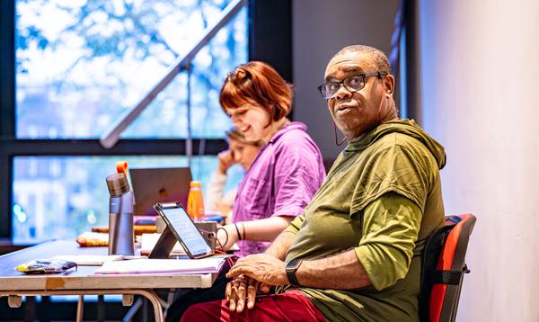 A photo of Clive Rowe and Clare Packham sat behind a desk in the rehearsal room. Clive Rowe is looking towards camera.