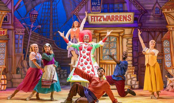 Clive Rowe as Sarah the Cook is wearing a dress of tomato sauce and is posing in the centre with arms in the air. The Ensemble are all posing around him, at different levels with their arms outstretched.