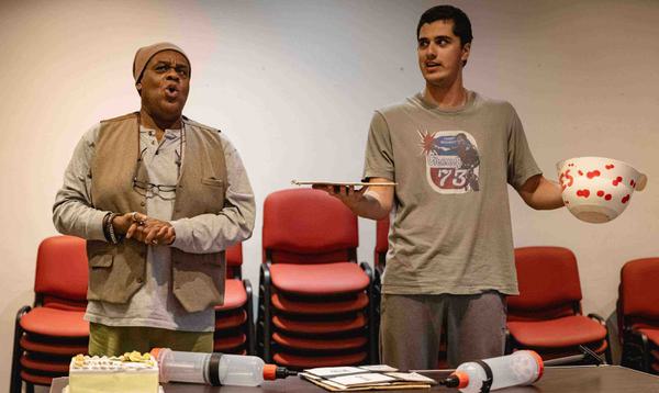 Clive Rowe as Sarah the Cook and Max Mirza as Idle Jack are positioned behind a table. Clive has a surprised look on his face and Max is holding a bowl and looks confused.