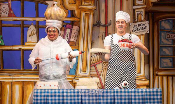 Clive Rowe as Sarah the Cook and Max Mirza as Idle Jack are stood behind a table holding cake icing, a cake and a bowl of cherries, with gleeful expressions on their faces.