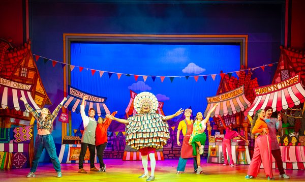 Clive Rowe as Widow Twankey is wearing a dress made out of fake money while the Ensemble pose around him in dance positions.