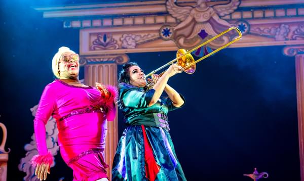 Clive Rowe as Widow Twankey is wearing a hot pink outfit and is following behind Natasha Lewis as Abby-na-zaaar! who is playing the trombone.