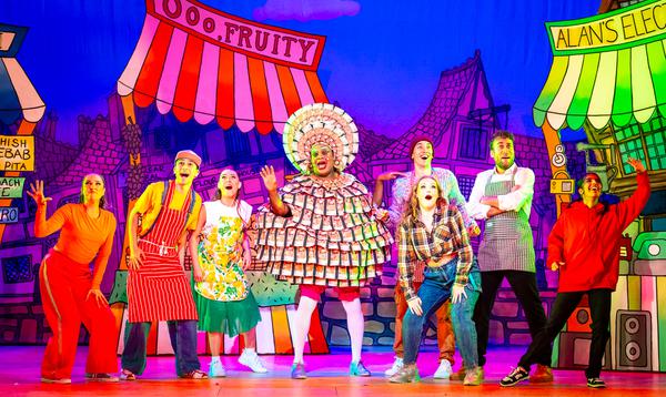 Clive Rowe as Widow Twankey is wearing a dress made out of fake money while the Ensemble pose around him looking excited.