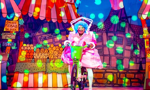 Clive Rowe as Widow Twankey is wearing a blue wig, a bright pink dress in the shape of a bag and is riding in on a Lime bike.