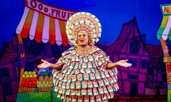 Clive Rowe as Widow Twankey is wearing a round dress made out of fake money and a headdress also made out of fake money and is gesturing outwards with his hands.