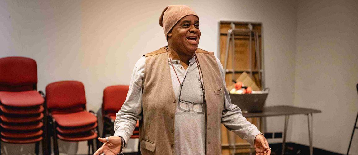 Clive Rowe as Sarah the Cook has both arms out low at the side of him and is smiling out front.