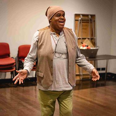 Clive Rowe as Sarah the Cook has both arms out low at the side of him and is smiling out front.
