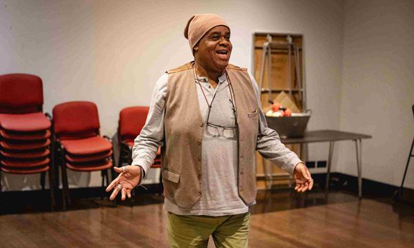 Clive Rowe as Sarah the Cook has both arms out low at the side of him and is smiling out front.