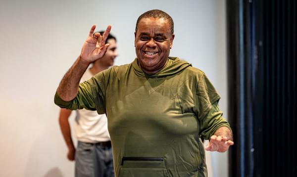 Clive Rowe is wearing a green jumper and is smiling with his hand in the air.