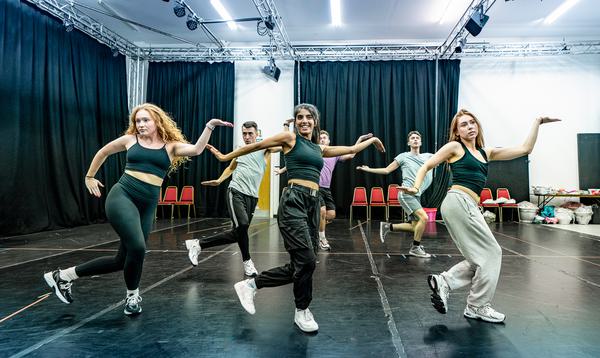 The Aladdin Ensemble are in the rehearsal room practicing a dance routine with their arms outstretched.