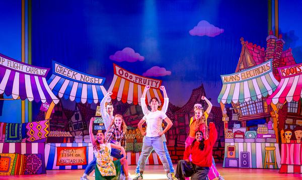 Fred Double as Aladdin is wearing a white t-shirt and jeans and is standing with hands on hips while the Ensemble pose around him.