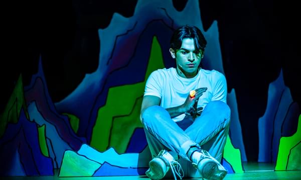 Fred Double as Aladdin is sitting on the ground in a greenish light and staring at the ring on his finger which is lit up.