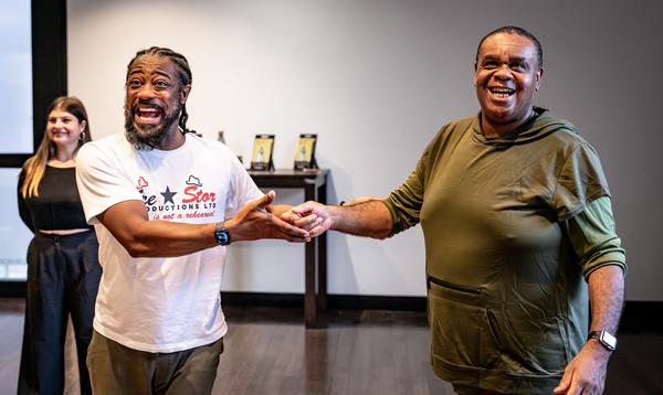 Ruth Lynch stands at the back with her hands behind her back whilst Kat B and Clive Rowe hold hands and smile towards the camera, in the middle of singing.