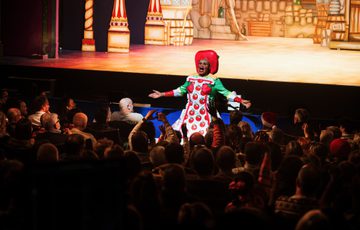 Clive Rowe as Sarah the Cook is stood at the front of the auditorium speaking with arms outstretched whilst the audience applaud.
