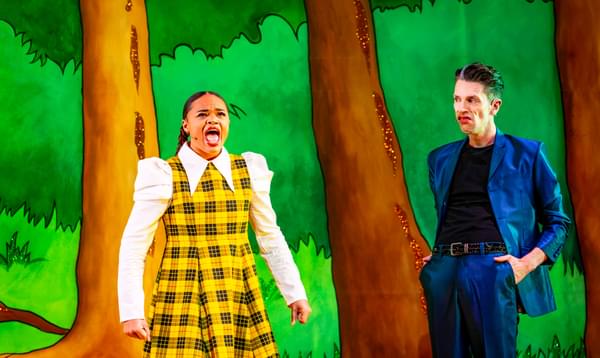 Isabella Mason as Jazz is wearing a yellow checked dress over a white blouse and looks very cross while George Heyworth as Mildew Funk looks on with a grimace on his face.