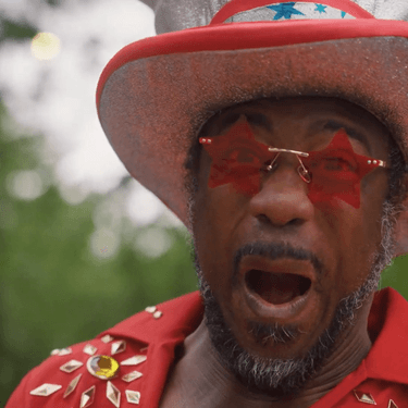 Kat B dressed in a red jumpsuit, large hat and red star sunglasses staring at camera with a shocked and excited expression, mouth open and eyes wide.