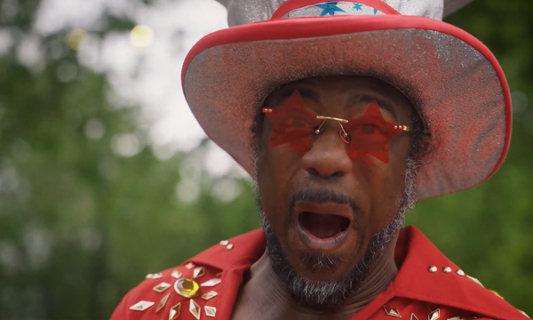 Kat B dressed in a red jumpsuit, large hat and red star sunglasses staring at camera with a shocked and excited expression, mouth open and eyes wide.