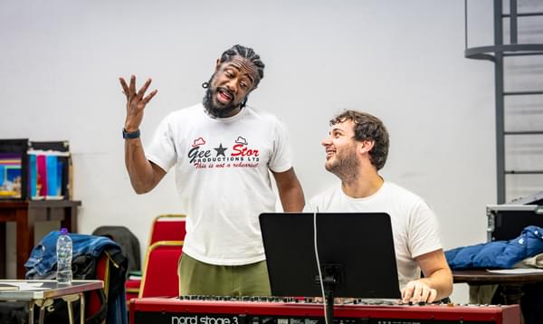 Kat B is singing with his hand in the air and is standing next to Musical Director Alex Maynard while he plays the keyboard.
