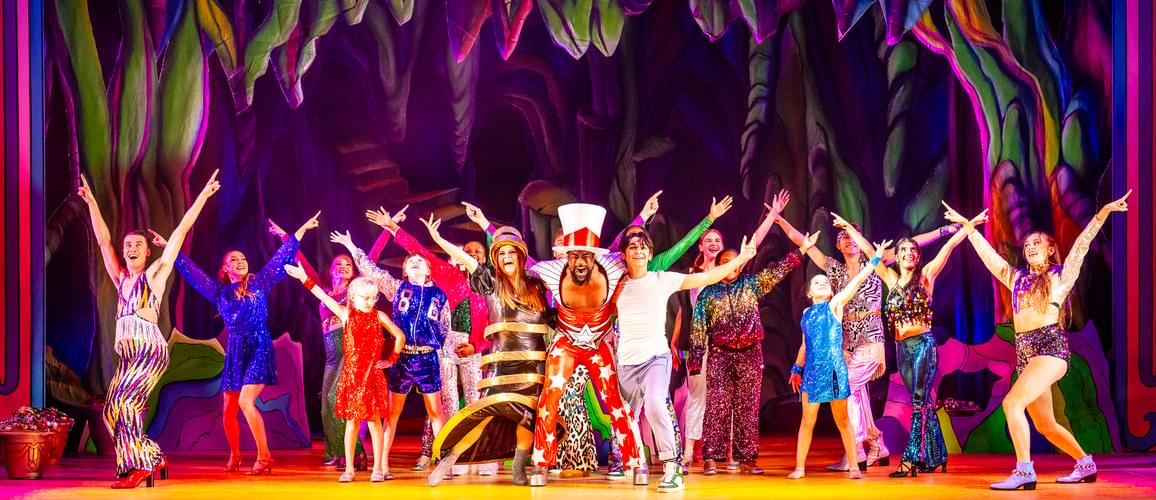 A photo of Kat B as the Genie wearing a red jumpsuit with Ruth Lynch as Spirit of the Ring and Fred Double as Aladdin either side and the full Ensemble and Young Ensemble posing around them.