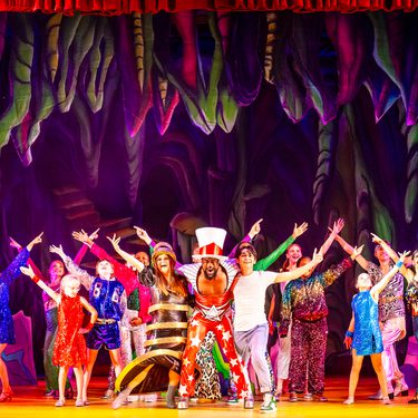 A photo of Kat B as the Genie wearing a red jumpsuit with Ruth Lynch as Spirit of the Ring and Fred Double as Aladdin either side and the full Ensemble and Young Ensemble posing around them.