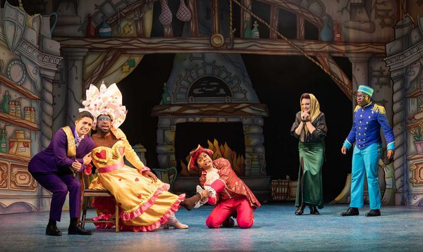 Luke Latchman as Prince Charming, Kat B as Flatula, Chrissie Bhima as Dandini, Alexandra Waite Roberts as Oblivia and Nicholas McLean as Buttons pose together with Kat B trying on the glass slipper and everyone else with pained/worried expressions.