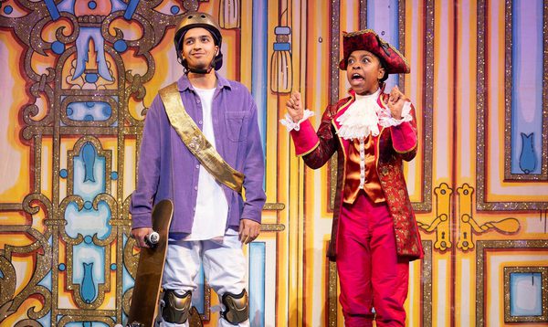 Luke Latchman as Prince Charming has a wry smile and is holding a skateboard and Chrissie Bhima as Dandini has her hands up and is speaking enthusiastically
