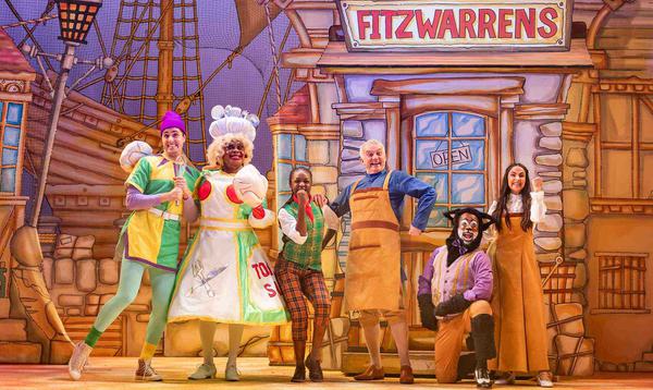 Max Mirza as Idle Jack, Clive Rowe as Sarah the Cook, Kandaka Moore as Dick Whittington, Tony Timberlake as Alderman Fitzwarren, Kat B as Tommy the Cat and Aryana Ramkhalawon as Alice Fitzwarren pose together in the middle of the stage, all looking happy and excited.