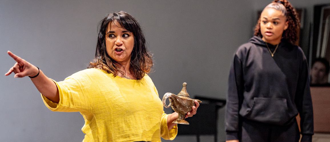 Natasha Lewis is wearing a yellow jumper with blue jeans and is holding a gold lamp while pointing to the right. Isabella Mason is standing beside her in a black jumper looking at her with a shocked expression.