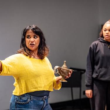 Natasha Lewis is wearing a yellow jumper with blue jeans and is holding a gold lamp while pointing to the right. Isabella Mason is standing beside her in a black jumper looking at her with a shocked expression.