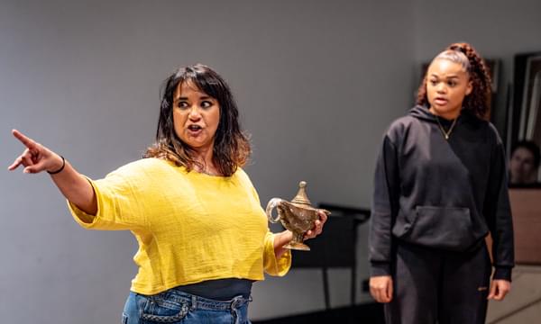 Natasha Lewis is wearing a yellow jumper with blue jeans and is holding a gold lamp while pointing to the right. Isabella Mason is standing beside her in a black jumper looking at her with a shocked expression.